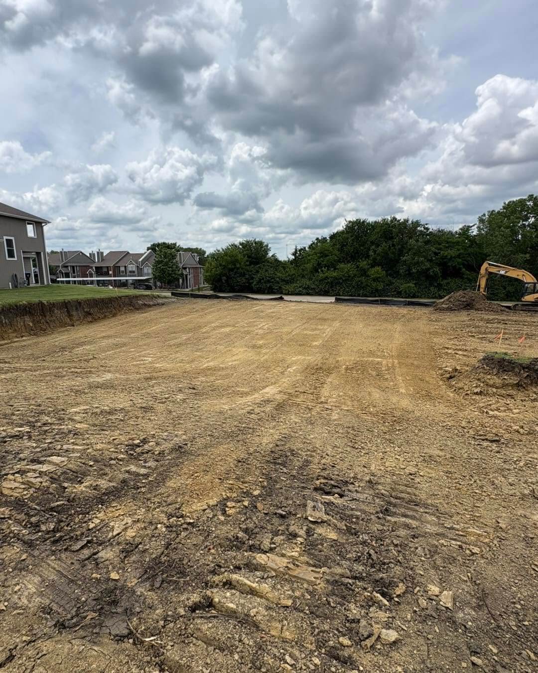 Cleared construction site with graded dirt, suburban homes, trees, and an excavator under dramatic storm clouds, suggesting new development.