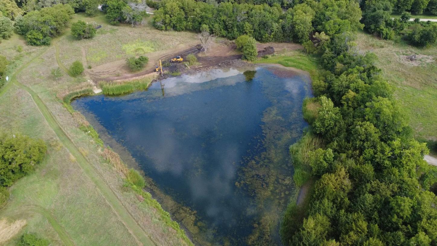 Aerial view of a pond surrounded by greenery, with a small area of land cleared and machinery visible near the edge.
