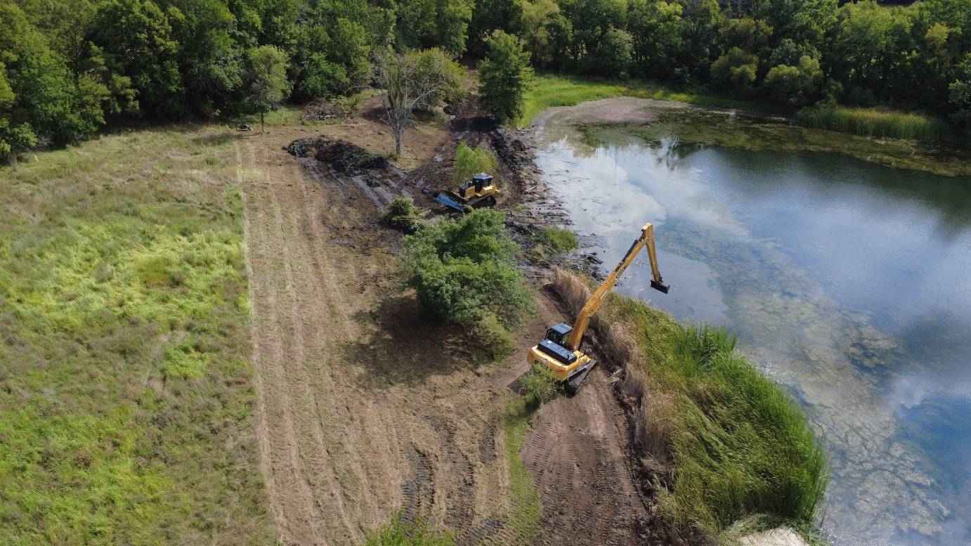 Aerial view of two yellow excavators reshaping a pond shoreline, with muddy banks, grassy fields, calm water, and dense green trees.