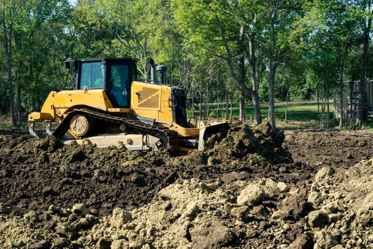 Yellow bulldozer pushes through freshly churned soil at a wooded construction site, surrounded by trees and dappled sunlight.