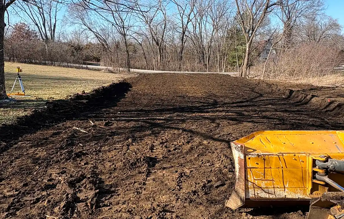 A cleared area of dirt with a yellow plow attachment in the foreground, surrounded by bare trees and a grassy field.