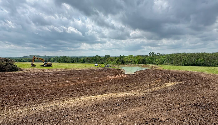 A large, open field with freshly disturbed soil and a small pond. Heavy machinery, including an excavator, is visible in the background under a cloudy sky.