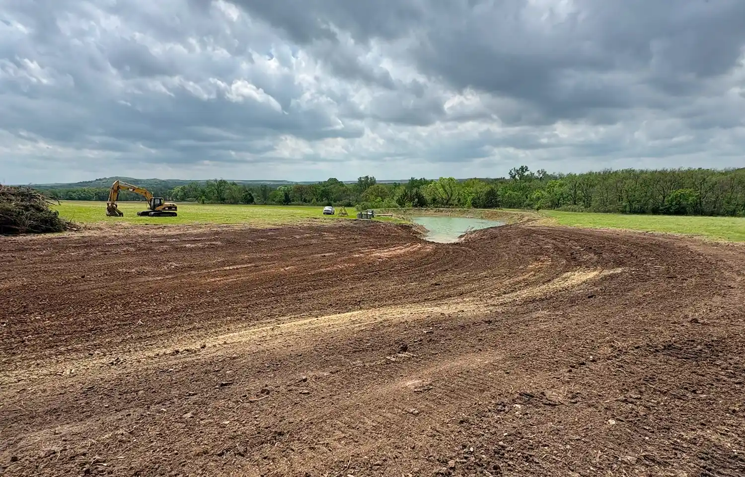 A large, open field with freshly disturbed soil and a small pond. Heavy machinery, including an excavator, is visible in the background under a cloudy sky.