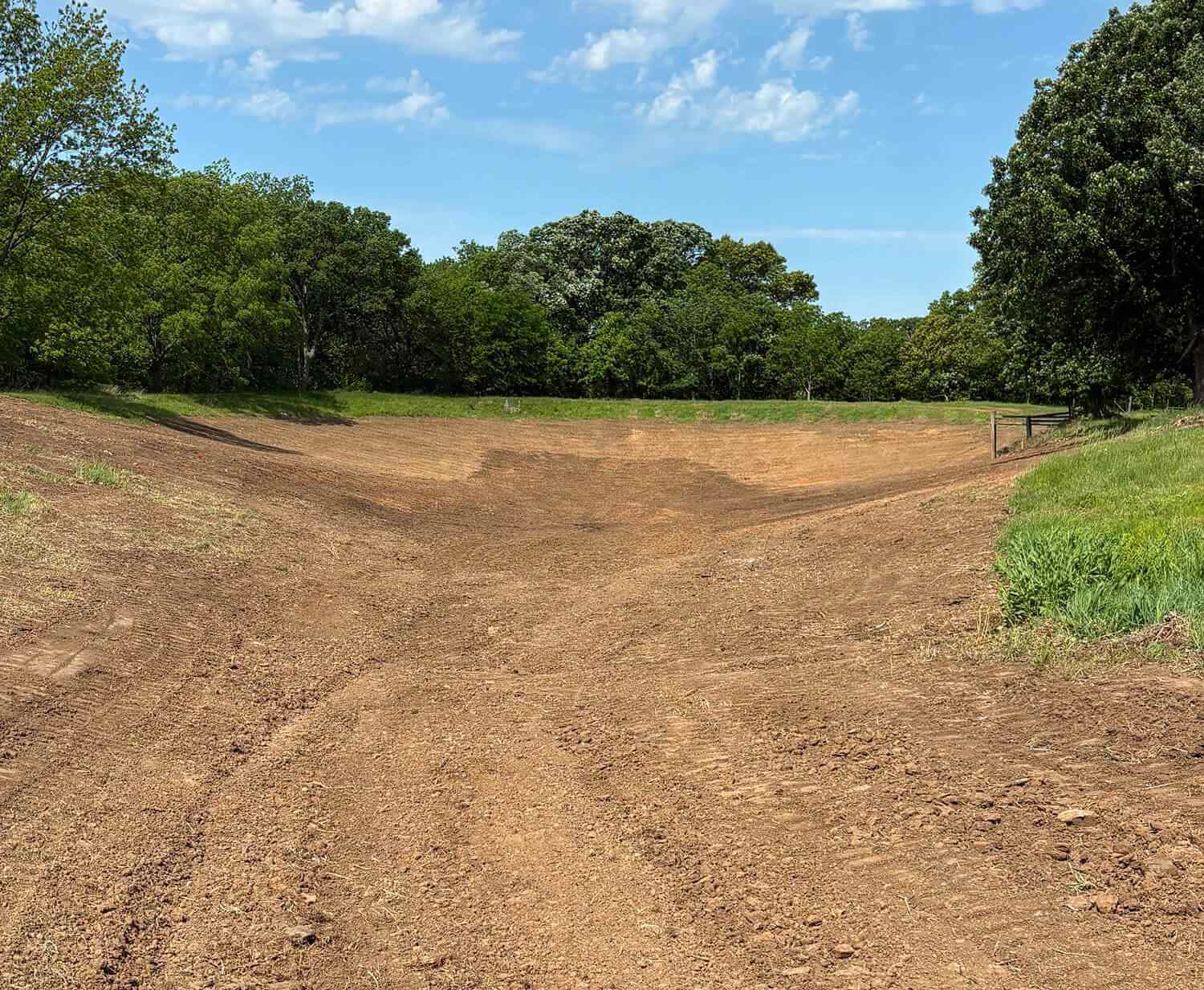 A broad, freshly tilled dirt field stretches toward a tree-lined horizon and wire fence under a bright blue sky, creating a quiet rural scene.