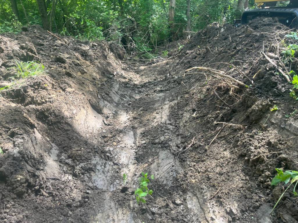 Deep muddy trench cuts through a lush forest, with exposed roots and soil; an off-road vehicle is partly visible at upper right.