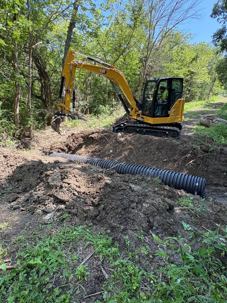 Yellow mini excavator digs a forest trench beside a long black corrugated drainage pipe, surrounded by trees and sunlit greenery.