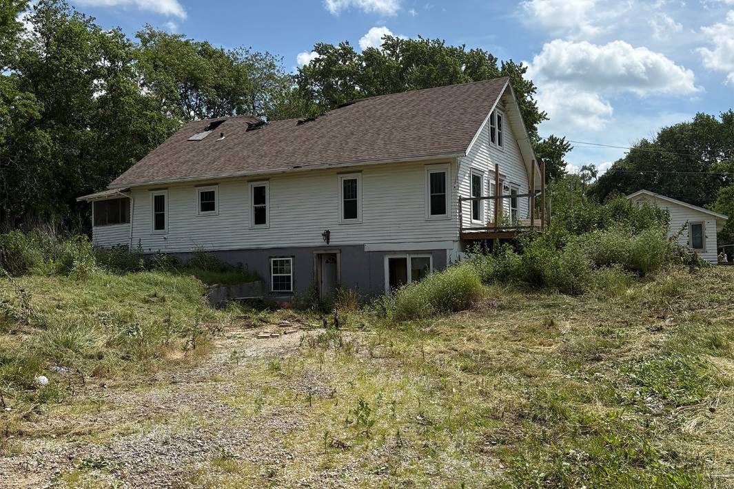 Weathered white farmhouse with a brown gabled roof and small deck sits in an overgrown yard beneath a bright blue sky, evoking quiet rural solitude.