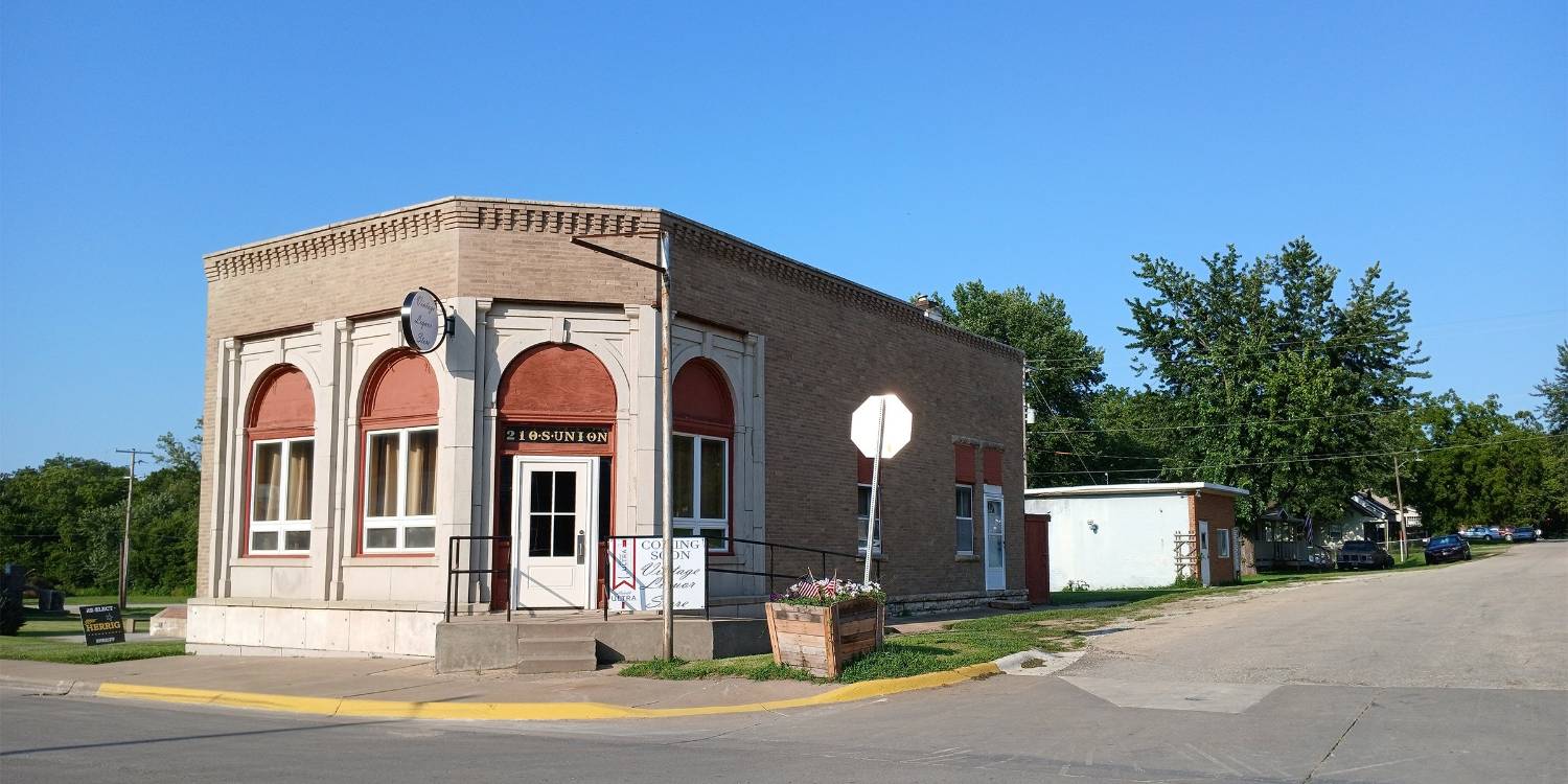 A small, beige building with arched windows and a flat roof, situated at a street corner with a stop sign and trees nearby.