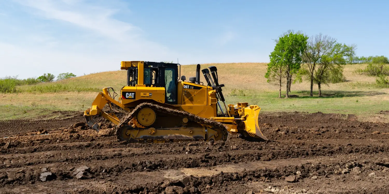 Yellow Caterpillar bulldozer grading dark soil in a rural field, with rolling hills and trees beneath blue skies; industrious, bright scene.