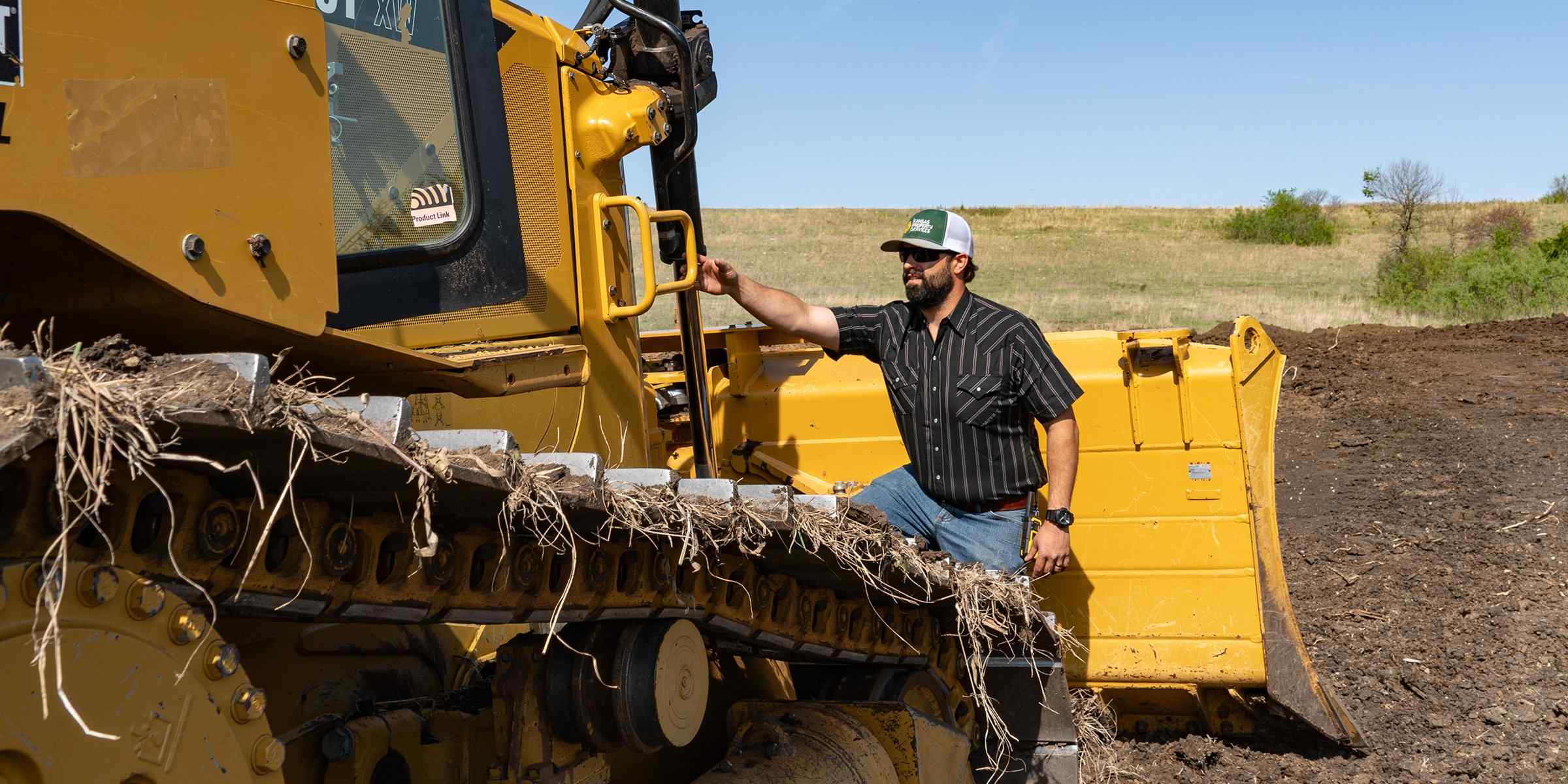 A man in a striped shirt and cap is standing next to a large yellow bulldozer in a field with a clear blue sky.