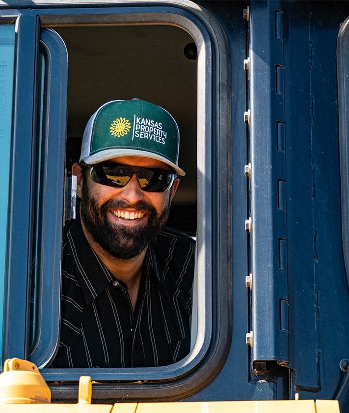 A man wearing sunglasses and a cap with the text "Kansas Property Services" smiles from the window of a vehicle.