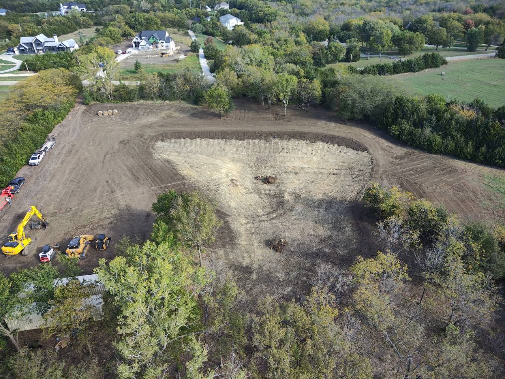 Aerial view of freshly tilled farmland curving around green fields and scattered trees, with a distant water tower creating a quiet rural scene.