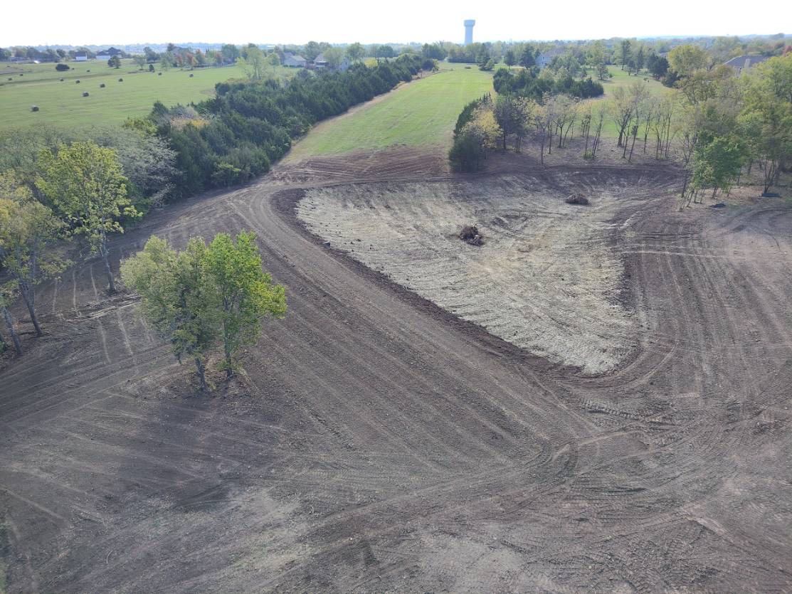 Aerial view of freshly tilled farmland curving around green fields and scattered trees, with a distant water tower creating a quiet rural scene.