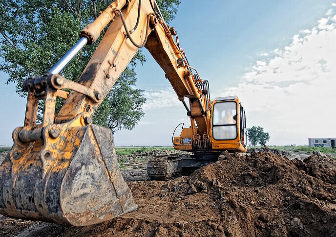 A yellow excavator is digging in a dirt field under a clear sky with a few trees and a distant building in the background.