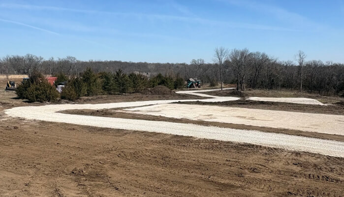 A construction site with a road curving through the landscape, featuring bare earth, trees in the background, and machinery in use.