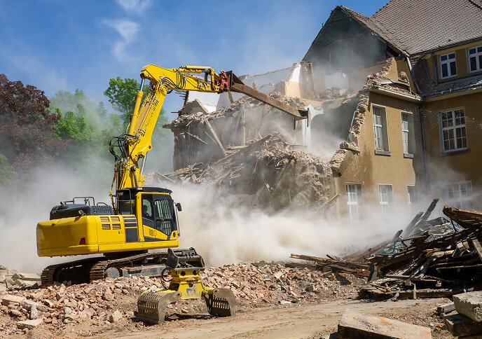 A yellow excavator demolishing a building, with debris and dust in the air.