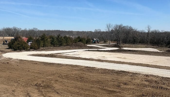 A construction site with a partially completed gravel road and surrounding trees under a clear blue sky.