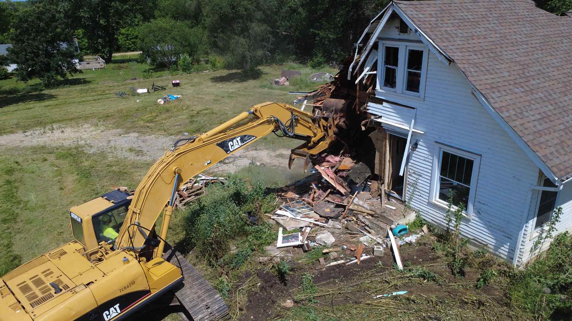 A large excavator demolishing the side of a white house, with debris scattered around and a rural landscape in the background.