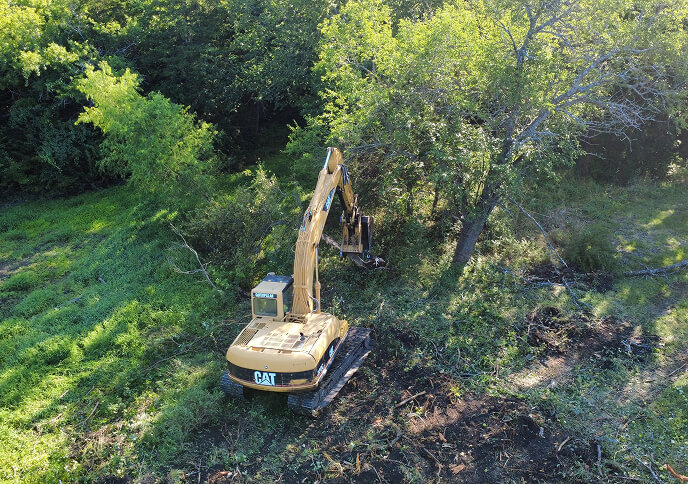 Aerial view of an excavator working in a green field, surrounded by trees and brush.