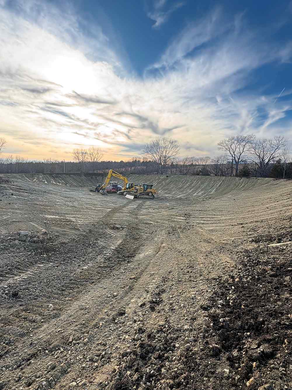 Yellow excavator and bulldozer sit on a barren construction site, surrounded by graded earth and leafless trees beneath a dramatic sky.