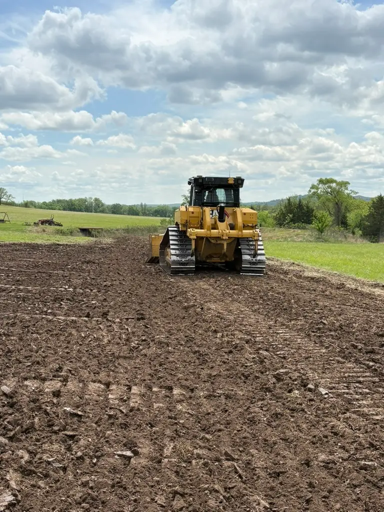Yellow Caterpillar bulldozer grading dark soil in a rural field, with rolling hills and trees beneath blue skies; industrious, bright scene.