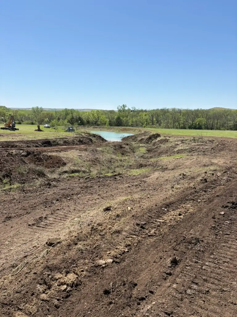 A large, open field with freshly disturbed soil and a small pond. Heavy machinery, including an excavator, is visible in the background under a cloudy sky.